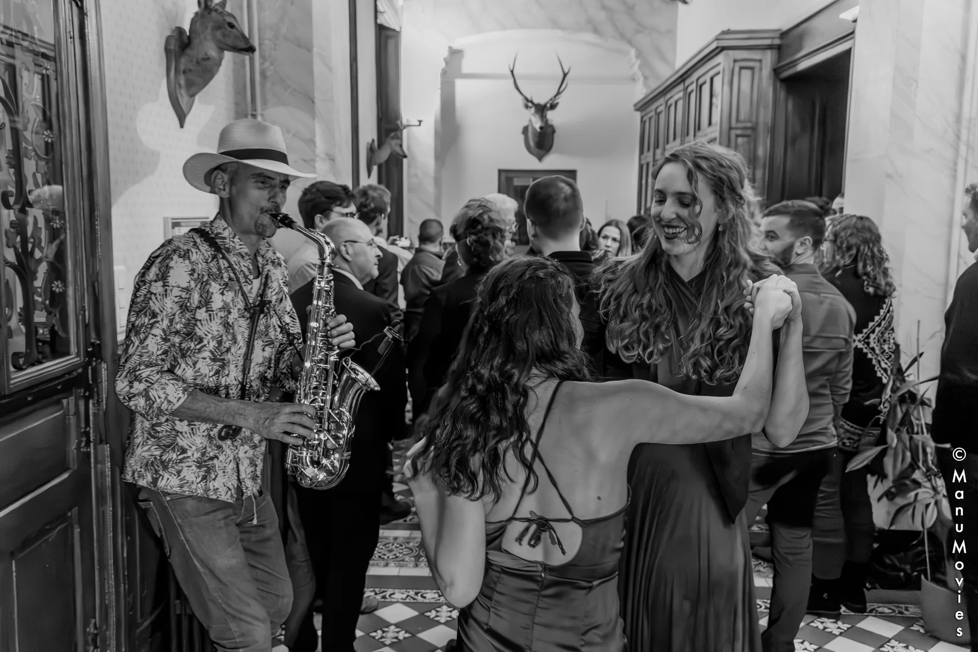 Saxophoniste premium pour vin d'honneur : mikOsax joue dans le hall d'un château pendant que les invités du mariage dansent (photo noir et blanc)